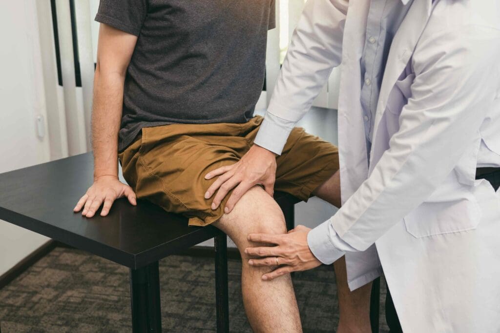 A bright, well-lit medical examination room with a middle-aged patient's lower body in the foreground, their knee joint exposed