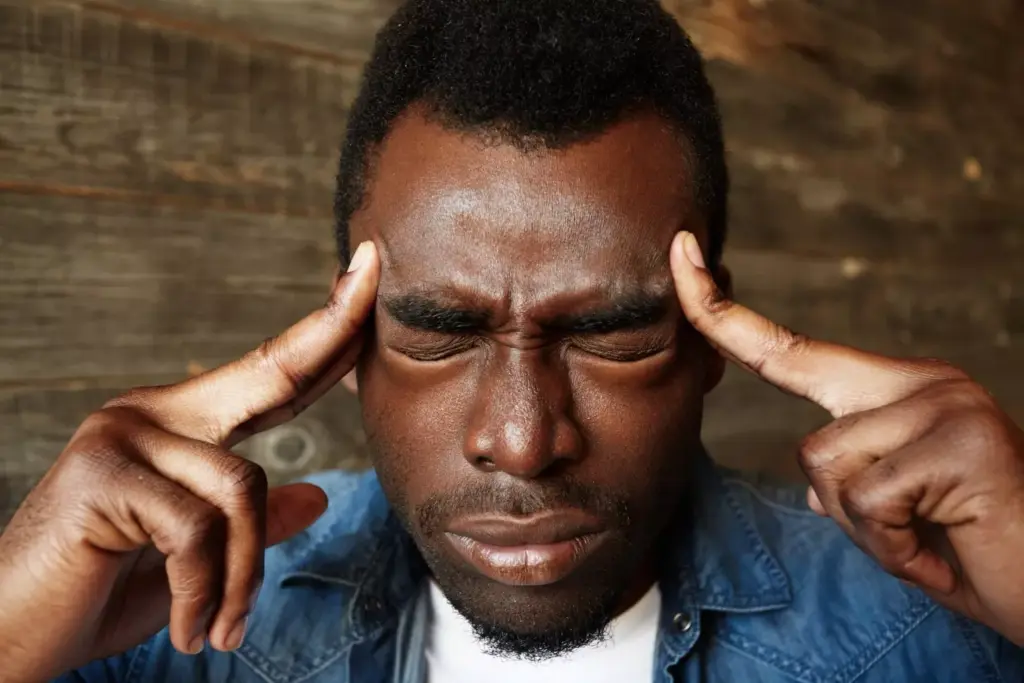 A close-up view of a human head, tilted to one side, with visible signs of distress on the face. The forehead is creased, the eyes are squinting, and the mouth is contorted in a grimace, conveying the intense pain of a persistent headache. The skin appears flushed, and the veins on the temples are visible, pulsing with each throb of the ache. The background is blurred, hazy, and dimly lit, creating a sense of isolation and the individual's sole focus on the debilitating discomfort. The lighting is soft and shadows are cast across the face, adding depth and dimension to the image. The overall composition and mood evoke the anguish and discomfort associated with a brain tumor-induced headache.