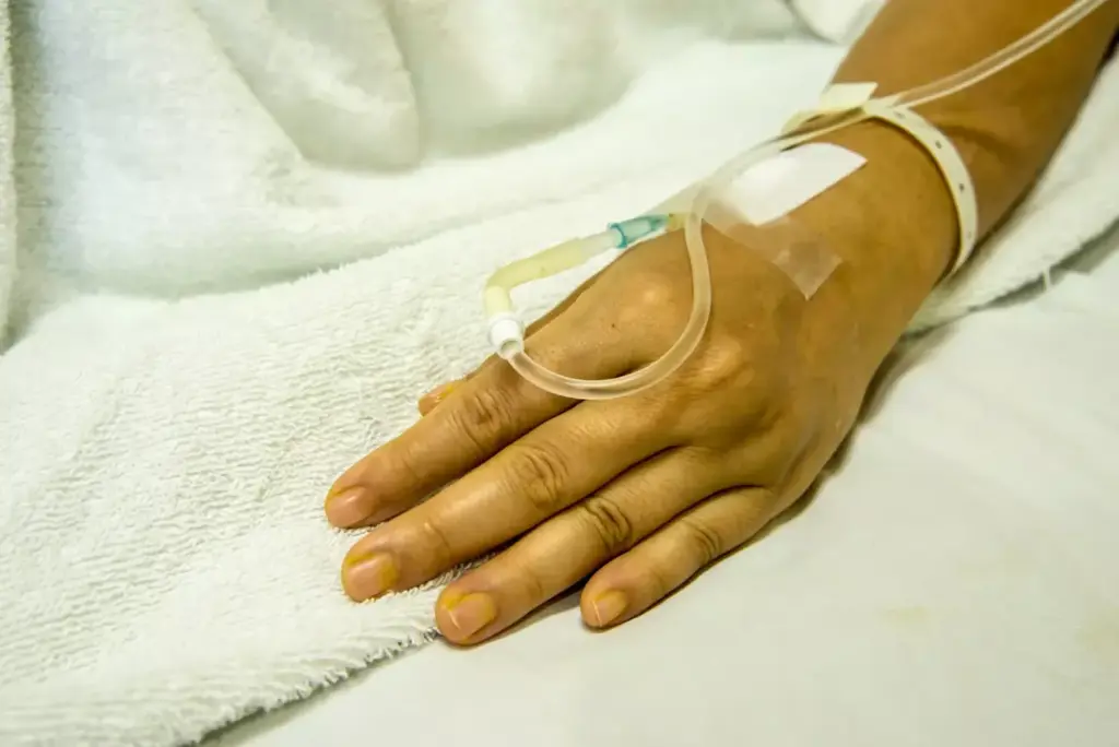 A close-up view of a patient's arm receiving chemotherapy treatment through an intravenous (IV) infusion. In the foreground, a clear plastic IV bag hangs from a metal stand, its contents slowly dripping into the patient's vein. The patient's arm is gently resting on a soft, supportive surface, with the IV line carefully taped in place. The background is blurred, focusing the viewer's attention on the precise, clinical process unfolding. Warm, natural lighting casts a comforting glow, while the overall atmosphere conveys a sense of care, professionalism, and the patient's well-being. The scene captures the essential elements of the chemo IV infusion procedure in a visually compelling, medically accurate manner.