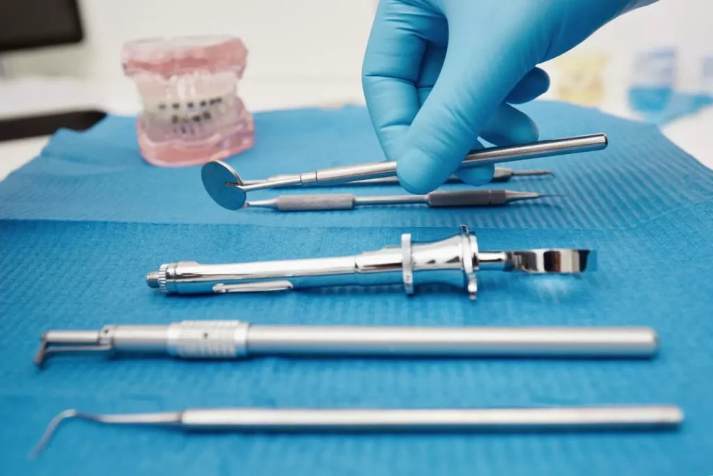 A close-up view of various dental bone graft materials neatly arranged on a clean, white surface. In the foreground, different shapes and sizes of bone graft particles, including xenografts, allografts, and synthetic bone substitutes, are displayed with clear separation between each type. The middle ground features surgical instruments like curettes and dental syringes, conveying the medical context. The background is softly blurred, maintaining the focus on the graft materials. Warm, directional lighting casts gentle shadows, highlighting the textural details of the grafts. The overall mood is one of clinical precision and medical expertise, suitable for illustrating the "Types of Dental Bone Graft Materials" section.