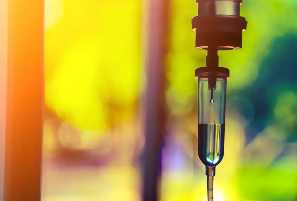 A detailed view of a chemotherapy IV drip process. In the foreground, a clear glass medical infusion bottle filled with a vibrant red liquid, hanging from a metal IV stand. The IV tube connects the bottle to a patient's arm, where the medication is steadily being administered. The middle ground features a blurred but recognizable medical environment, with medical equipment and personnel visible in the background. The lighting is soft, diffused, and clinical, creating a soothing, professional atmosphere. The camera angle is slightly elevated, providing a comprehensive view of the entire setup, emphasizing the technical precision and care involved in the chemotherapy infusion process. A detailed view of a chemotherapy IV drip process. In the foreground, a clear glass medical infusion bottle filled with a vibrant red liquid, hanging from a metal IV stand. The IV tube connects the bottle to a patient's arm, where the medication is steadily being administered. The middle ground features a blurred but recognizable medical environment, with medical equipment and personnel visible in the background. The lighting is soft, diffused, and clinical, creating a soothing, professional atmosphere. The camera angle is slightly elevated, providing a comprehensive view of the entire setup, emphasizing the technical precision and care involved in the chemotherapy infusion process.