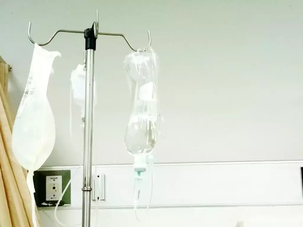 A minimalist, clinical depiction of the chemo IV infusion process. In the foreground, a clear intravenous bag hangs from a metal pole, the chemotherapy medication slowly dripping into a thin tube leading to the patient's arm. The middle ground shows the patient's hand resting comfortably, the needle skillfully inserted. The background blurs into a plain, sterile room, with soft, even lighting illuminating the scene. The overall mood is one of professionalism and medical precision, conveying the science-based nature of this cancer treatment.