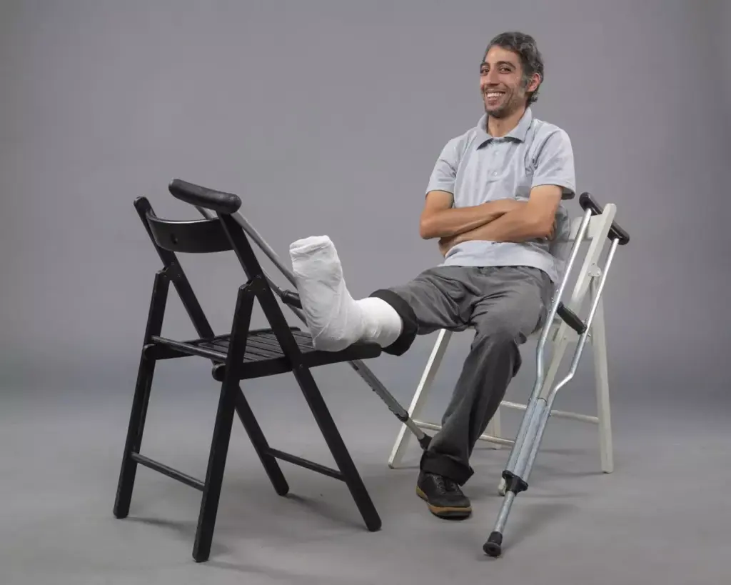 A patient sitting comfortably on a chair, leg propped up on a stool, with a knee brace visible. The background is a modern, light-filled physical therapy clinic, with clean white walls and equipment visible. The lighting is soft and diffuse, creating a serene and calming atmosphere. The subject's expression conveys a sense of cautious optimism as they begin their knee replacement recovery journey. The scene is captured using a wide-angle lens, providing a comprehensive view of the recovery process.