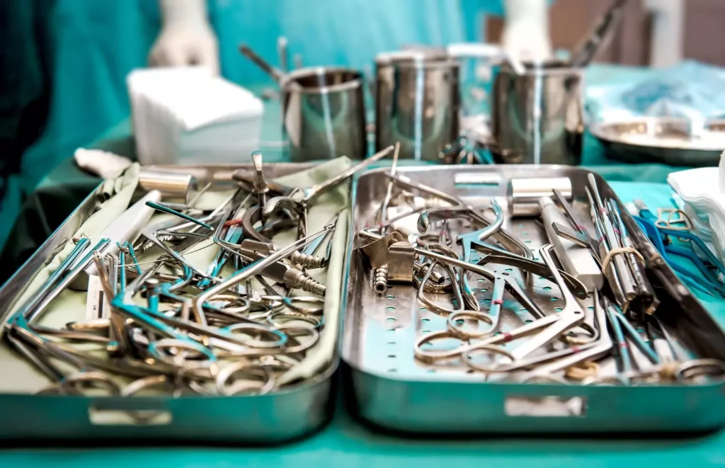 A still life photograph of various bone grafting materials used in dental implant procedures. In the foreground, a selection of synthetic bone graft granules, cortical bone chips, and demineralized bone matrix blocks. In the middle ground, surgical tools such as a dental curette, bone graft carrier, and bone tamp. The background is softly blurred, creating a clinical, medical atmosphere with muted gray tones and minimal distractions. Bright, even lighting from the top and sides highlights the textures and details of the materials. Shot with a macro lens at a shallow depth of field to draw the viewer's focus to the bone grafting components.