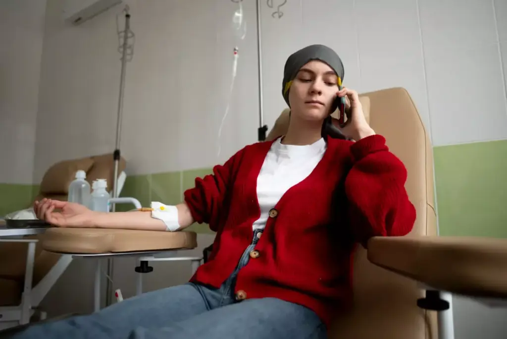 A woman sitting in a hospital chair, her head wrapped in a scarf, receiving an intravenous chemotherapy infusion. The room is softly lit, with muted tones of white, gray, and blue. The woman's expression is somber, yet resolute, as she gazes out a nearby window. Medical equipment, such as an IV stand and monitoring devices, are present, conveying the clinical setting. The background is blurred, creating a sense of focus on the woman and her treatment. The overall mood is one of quiet reflection, highlighting the emotional and physical challenges of breast cancer chemotherapy.